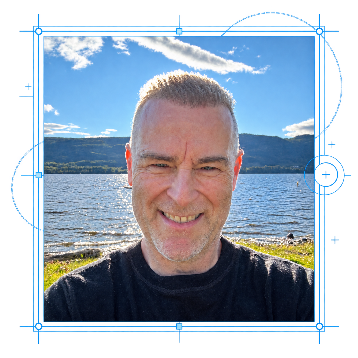 Smiling gray-haired man in a black shirt poses by a sunlit lake with hills and clouds, surrounded by white editing guides.
